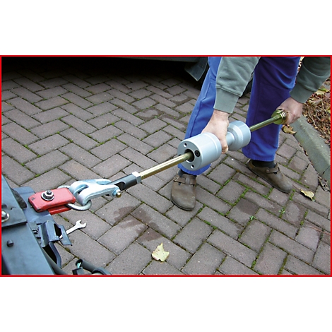 Eine Person hält ein Werkzeug, das an einer Anhängerkupplung befestigt ist. Der Hintergrund ist ein Pflastersteinweg.